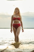 A woman with long hair walks confidently in shallow water, wearing a red bikini. The sun sets in