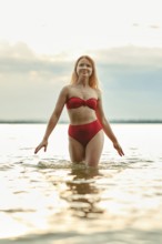 A young woman with long hair walks through shallow water, showcasing a vibrant red swimsuit. The