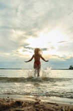 A woman in red swimwear plays in the water, splashing joyfully as the sun sets in the background.