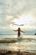 A woman in a red swimsuit wades into a shallow lake, arms outstretched, as the sun sets behind her.