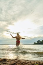 A woman in a red swimsuit plays in the shallow water of a lake. The sun sets behind her, casting a