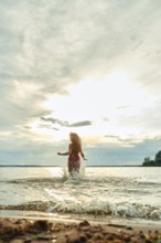 A woman with long hair playfully splashes in the water at a beach during sunset. The sky is