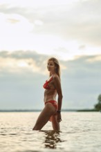 A woman in a red bikini walks through calm waters, silhouetted against a sunset with clouds