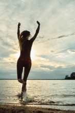 A woman runs through shallow water, joyfully throwing up her arms towards the setting sun