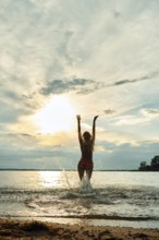 A woman stands in shallow water at a beach during sunset, joyfully raising her arms towards the