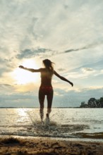 A woman wearing swimwear joyfully splashes through water at sunset. The sky displays vibrant