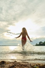 A young woman in a red swimsuit joyfully runs into the lake, creating splashes as she embraces the