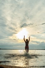 A person stands in the shallow water with arms raised, enjoying the sunset. The sky is filled with
