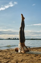 A woman performs shoulder blade stand with her legs raised high at a lakeside on a sandy shore. The