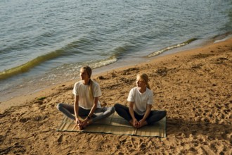 Two women are sitting on a mat by the water's edge and doing yoga poses as the sun begins to set.