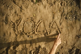 A person draws the word yoga in the sand using her finger. The setting is a sunny beach, showcasing