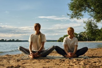 Two women in comfortable clothing are practicing yoga on the beach by a tranquil lake. They sit