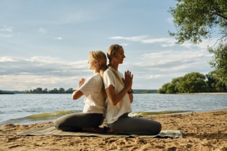 Two women sit back-to-back on a sandy lakeshore, practicing yoga under the warm evening sun. They