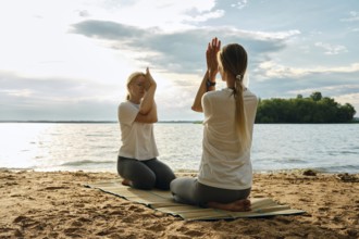 Two women are engaging in a yoga session on a sandy beach beside a tranquil lake