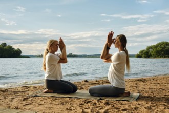 Two individuals are engaged in a yoga session on a sandy beach by a tranquil lake. The sun sets,