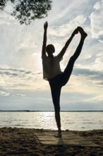 A woman balances gracefully on one leg while practicing yoga on a beach at sunset. The sun sets