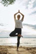 A woman performs yoga on a sandy beach, balancing in a tree pose with arms raised towards the sky.