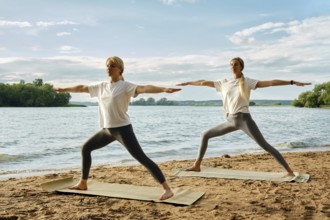 Two women perform yoga warrior pose on mats at a sandy beach by a calm lake. The sun is shining