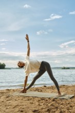 A person practices yoga on a mat by the water's edge during sunset. Woman stands in a triangle