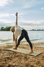 A woman stretches into a side angle pose on a yoga mat placed on the sandy shoreline