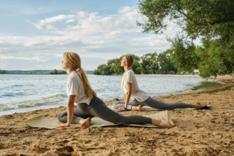 Two women engage in yoga poses on the sandy beach beside a calm lake. The sun shines brightly,