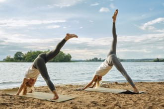 Two women practice yoga on mats along a sandy lakeside during the early evening. They rest their
