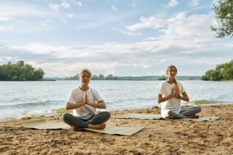 Two women are sitting cross-legged on yoga mats by the riverbank, practicing yoga with serene