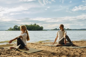 Two barefoot women practice yoga on mats on a sandy lakeshore at sunset