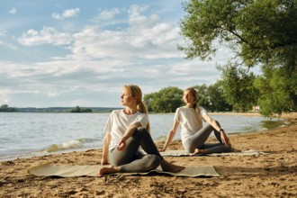 Two women practice yoga on a sandy lakeshore, surrounded by the calm waters and lush greenery. The