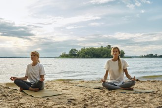 Two women sit cross-legged on yoga mats, meditating by a lake. Soft waves lap against the sandy