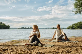 Two women practicing yoga on sandy shores by a river. Surrounded by trees and under a bright sky,