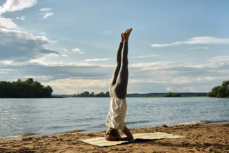 A woman performs a headstand yoga pose on a sandy beach beside a tranquil river. The sun sets in