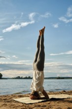 A woman balances in a headstand on a yoga mat on the sandy shoreline of a lake during sunset. The