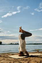 A slender woman performs a headstand yoga pose on a mat in the sand near a tranquil lake. The