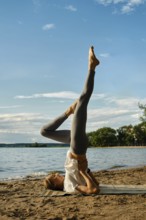 A dedicated individual performs a yoga pose on a sandy beach by the tranquil lake. The sun shines