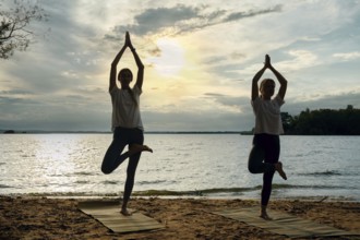 Silhouettes of two women performing yoga poses on mats along a sandy lakeshore as the sun sets,