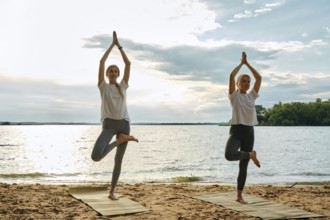 Two women perform yoga poses on mats at the beach during sunset. They are surrounded by a tranquil