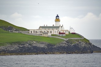 A lighthouse on a green coastal cliff next to the sea under a cloudy sky, Shetland Islands,