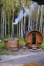 A cozy wooden sauna and hot tub surrounded by tall birch trees. Smoke gently rises from the