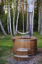 A rustic wooden hot tub sits in a peaceful clearing, surrounded by tall trees and green grass. A