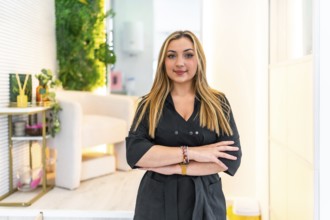 Young blond female beautician smiling with arms crossed in her beauty salon, vertical portrait
