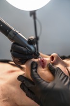 Beautician wearing black gloves applying permanent makeup on lips using a tattoo machine in a