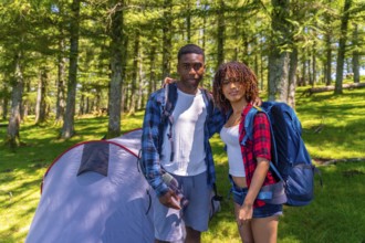 Young couple of hikers embracing and smiling next to their tent in a lush forest, enjoying a joyful