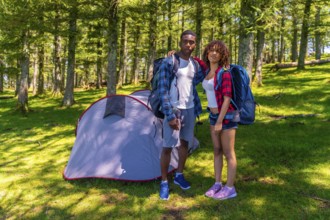 Young hikers posing together next to their tent in a beautiful sunny forest, enjoying the