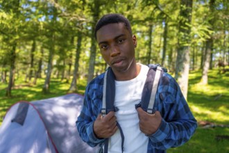 Young black man adjusts his backpack straps while standing in a forest clearing near his tent,