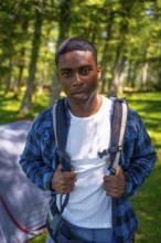 Young black man is wearing a backpack and standing in a forest near his tent, enjoying the peace of