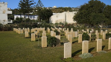 An extensive war cemetery with gravestones, surrounded by trees and a hilly landscape under a blue
