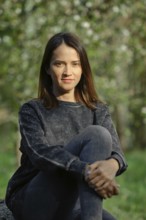 A young woman with long dark hair sits comfortably on a rock, wearing a casual sweater and leggings