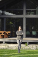 A young woman stands crossing her hands on the lawn in front of her modern house with large glass