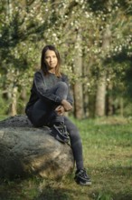 A young woman with long hair sits thoughtfully on a large rock in a peaceful forest. She wears